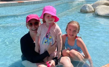 A woman and two children in a pool, wearing pink hats and swimsuits, smiling on a sunny day.