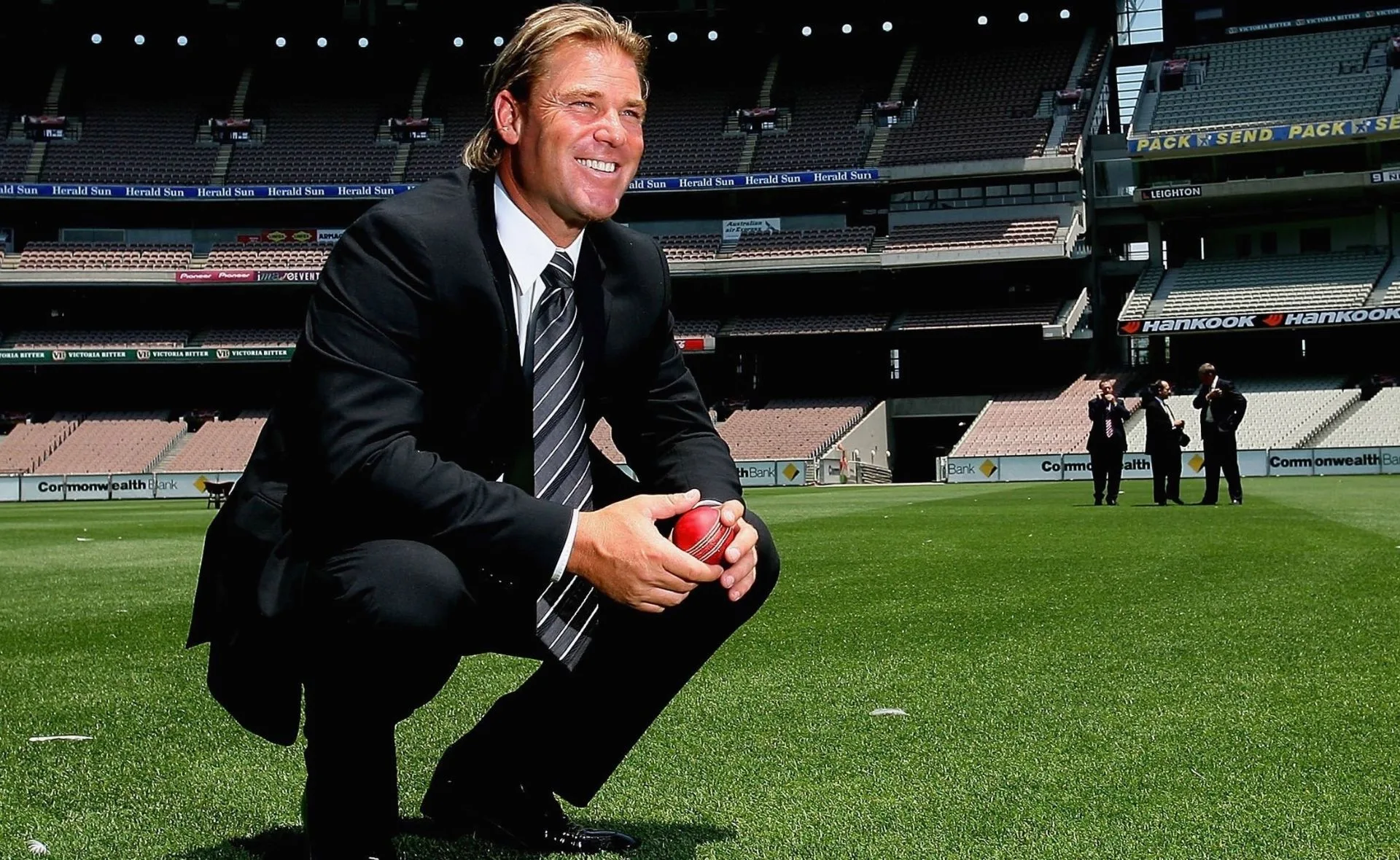Shane Warne in a suit on a cricket field, holding a red ball, with stadium seating in the background.