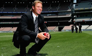 Shane Warne in a suit on a cricket field, holding a red ball, with stadium seating in the background.