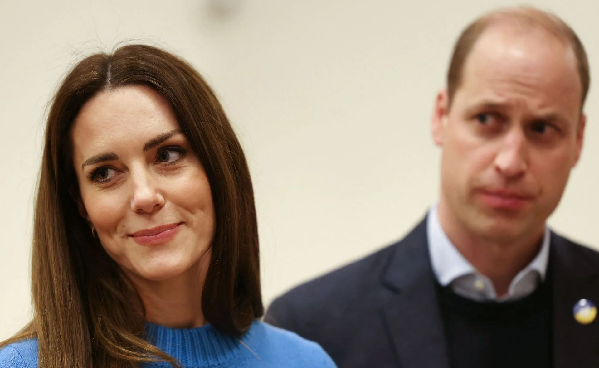 A woman in a blue sweater smiles slightly, with a man beside her looking ahead intently.