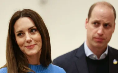 A woman in a blue sweater smiles slightly, with a man beside her looking ahead intently.