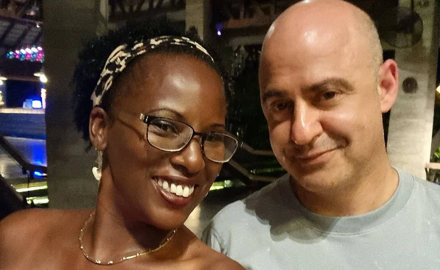 A woman and man smiling, posing for a selfie indoors with soft lighting in the background.