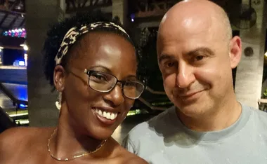 A woman and man smiling, posing for a selfie indoors with soft lighting in the background.