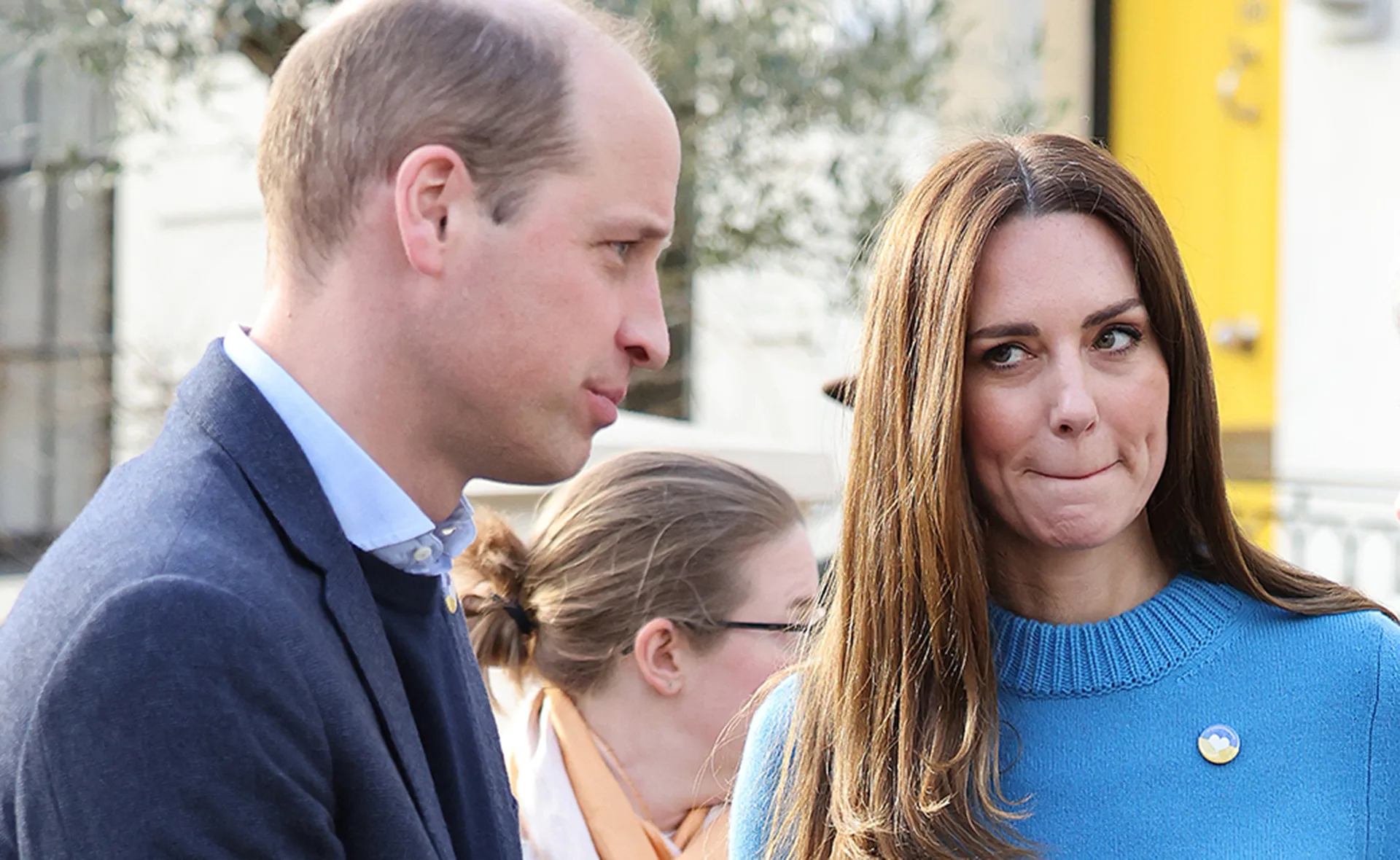 Two people are talking outside; one wears a blue sweater with a pin, the other a blue jacket. A third person is in the background.
