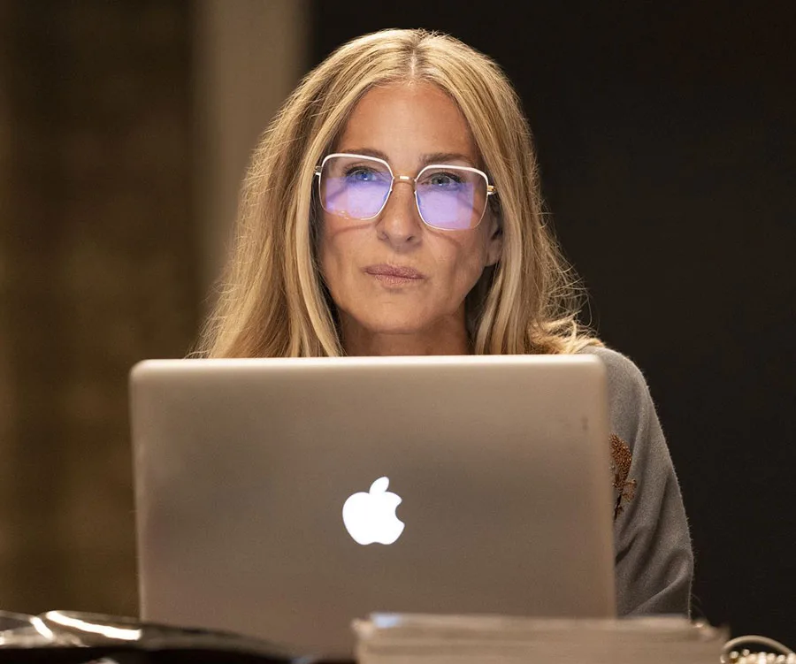 Woman with long hair and blue-tinted glasses working on a laptop, focused expression, stacks of paper nearby.
