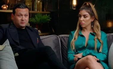 A man and woman sitting on a couch, engaged in a conversation, with a neutral background setting.