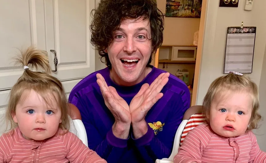 Man in a purple shirt smiling with two young children in matching pink outfits seated beside him.