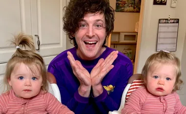 Man in a purple shirt smiling with two young children in matching pink outfits seated beside him.