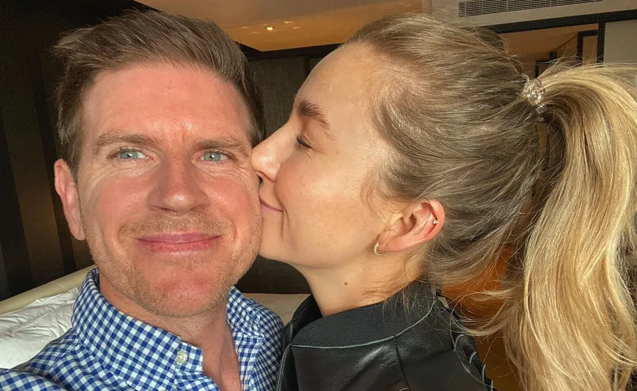 Man smiling as a woman kisses his cheek, both look happy, indoors setting.