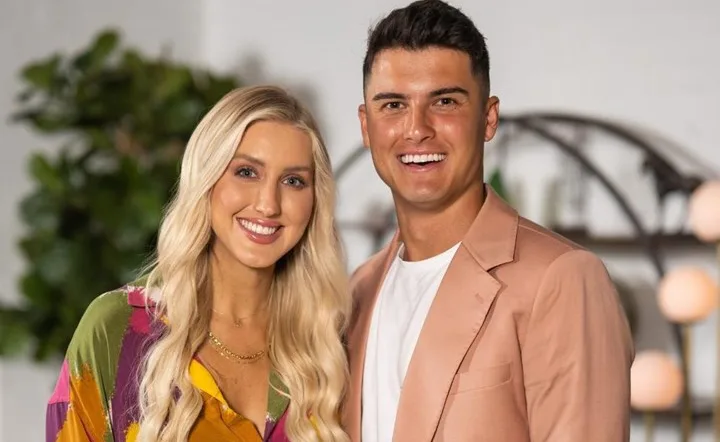 Couple smiling together, with the woman in a colorful dress and the man in a peach jacket, standing indoors.