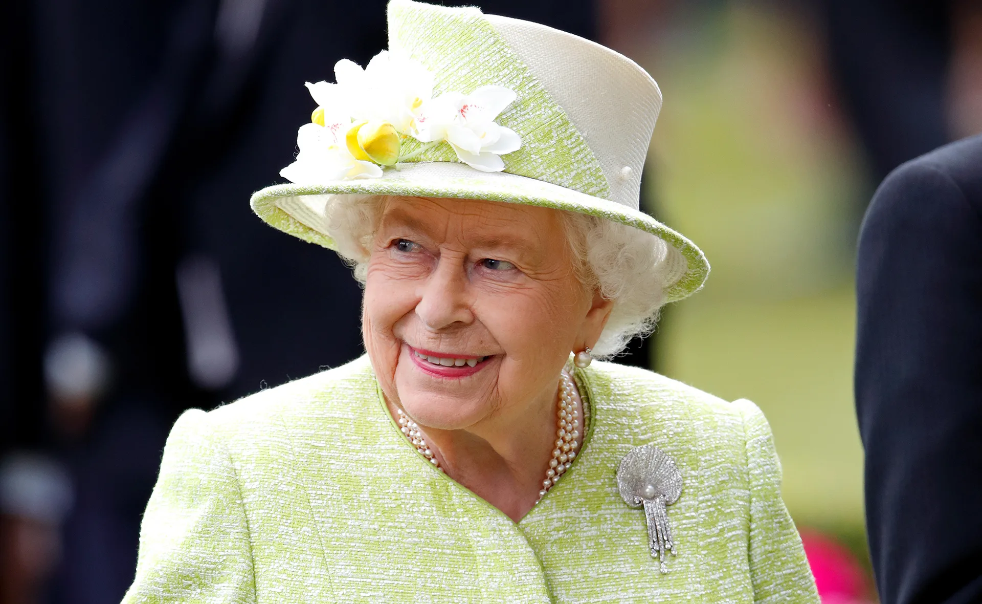 Elderly woman in a light green outfit and hat with floral decoration, smiling outdoors.