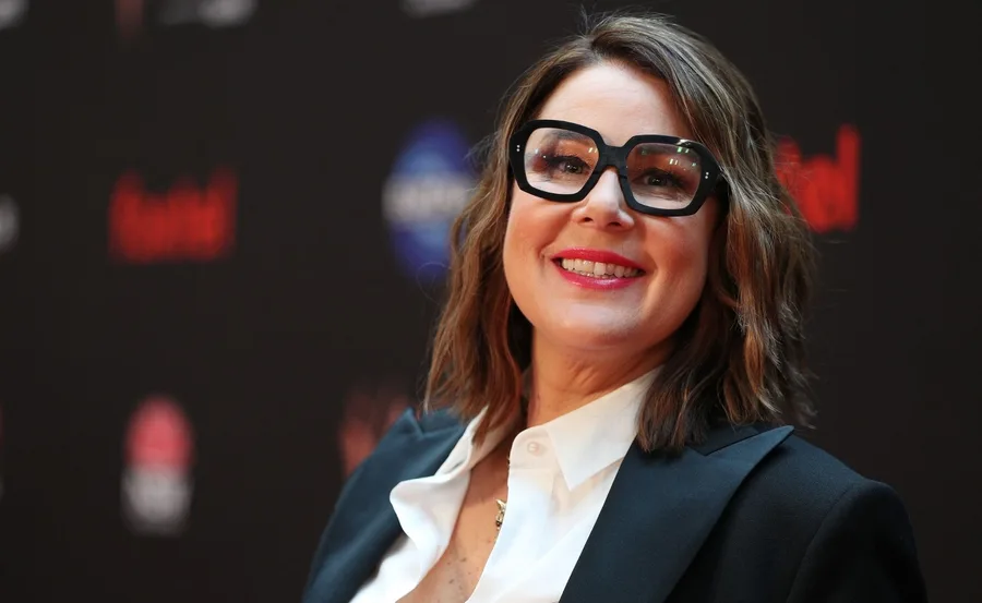 A person with shoulder-length hair and glasses smiles, wearing a black blazer and white shirt, on a dark background.