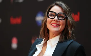 A person with shoulder-length hair and glasses smiles, wearing a black blazer and white shirt, on a dark background.