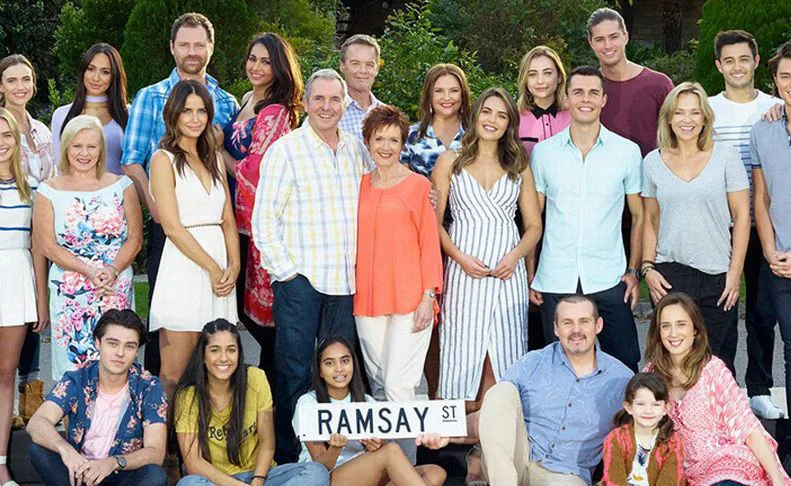 A large group of people stands together outdoors, holding a "Ramsay St" sign, smiling for a group photo.