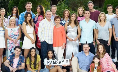A large group of people stands together outdoors, holding a "Ramsay St" sign, smiling for a group photo.