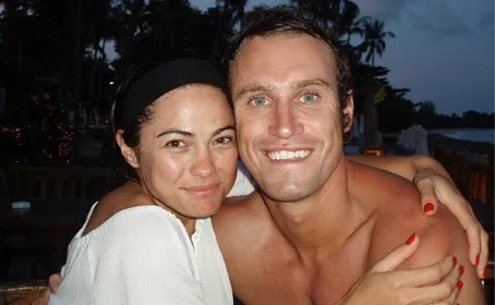 A couple smiling widely and embracing, sitting outdoors with a tropical evening background.