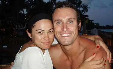 A couple smiling widely and embracing, sitting outdoors with a tropical evening background.