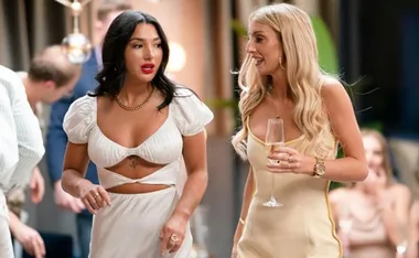 Two people at a party, one in a white dress and the other in a champagne-colored dress, chatting and holding drinks.