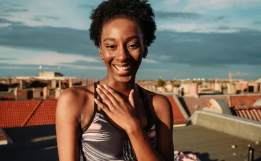 Smiling woman in activewear stands on a rooftop, hand on chest, with a cityscape background under a partly cloudy sky.