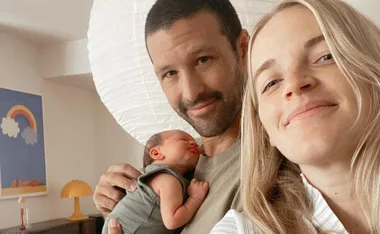 A couple smiling at the camera, holding a newborn baby. A colorful rainbow painting is in the background.