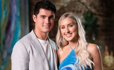 A man in a light gray suit and a woman in a blue dress smile together in a warmly lit room.
