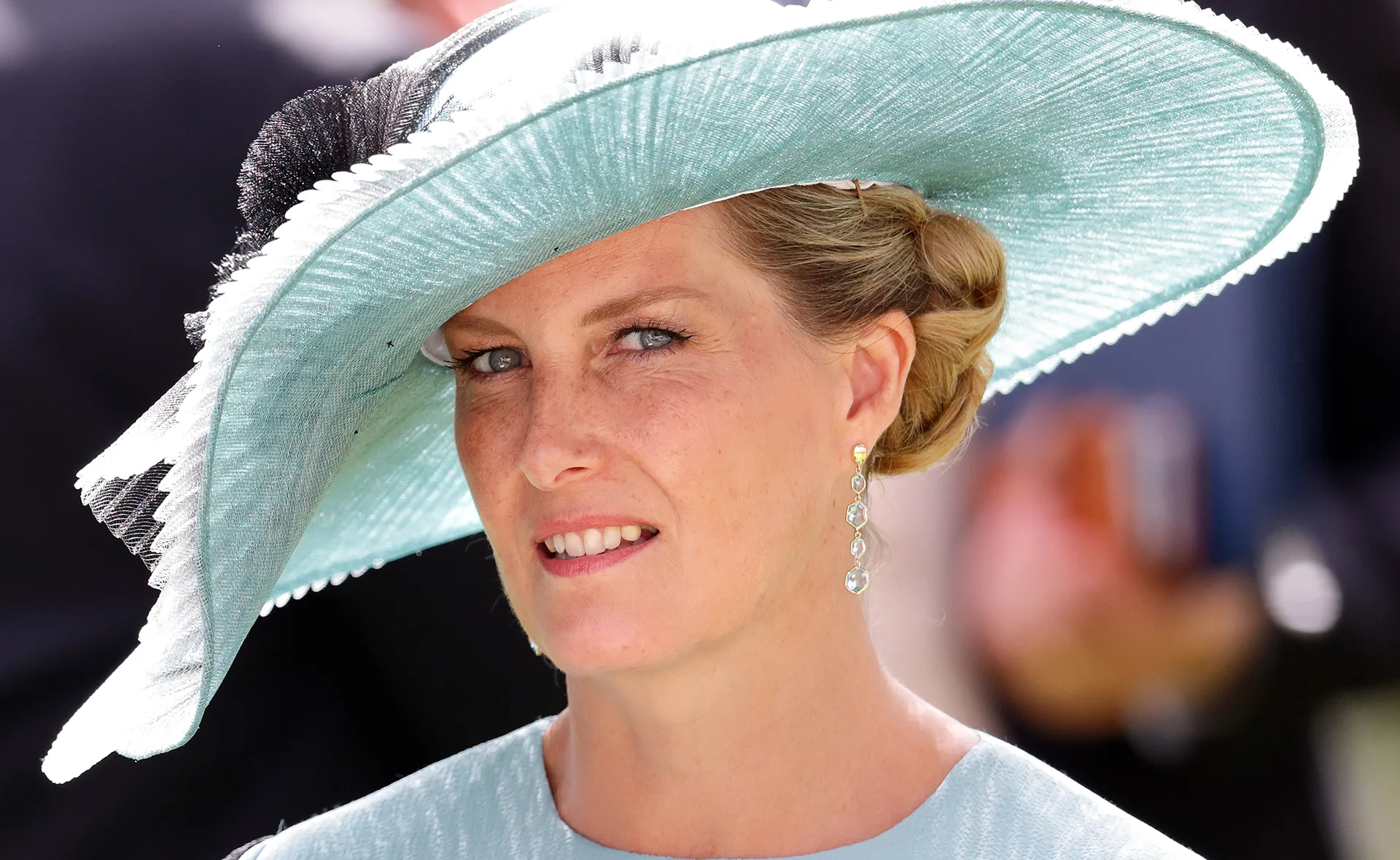 Woman in a large, light blue hat with elegant earrings attending an event outdoors.