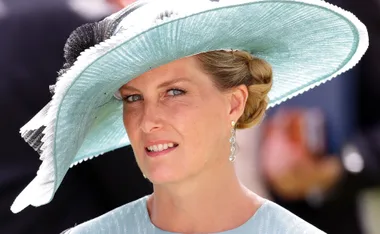 Woman in a large, light blue hat with elegant earrings attending an event outdoors.