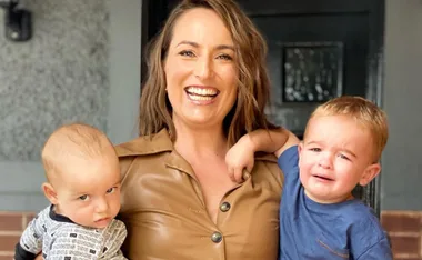 A woman smiling, holding two young children, one looking curious and the other crying, standing in front of a door.