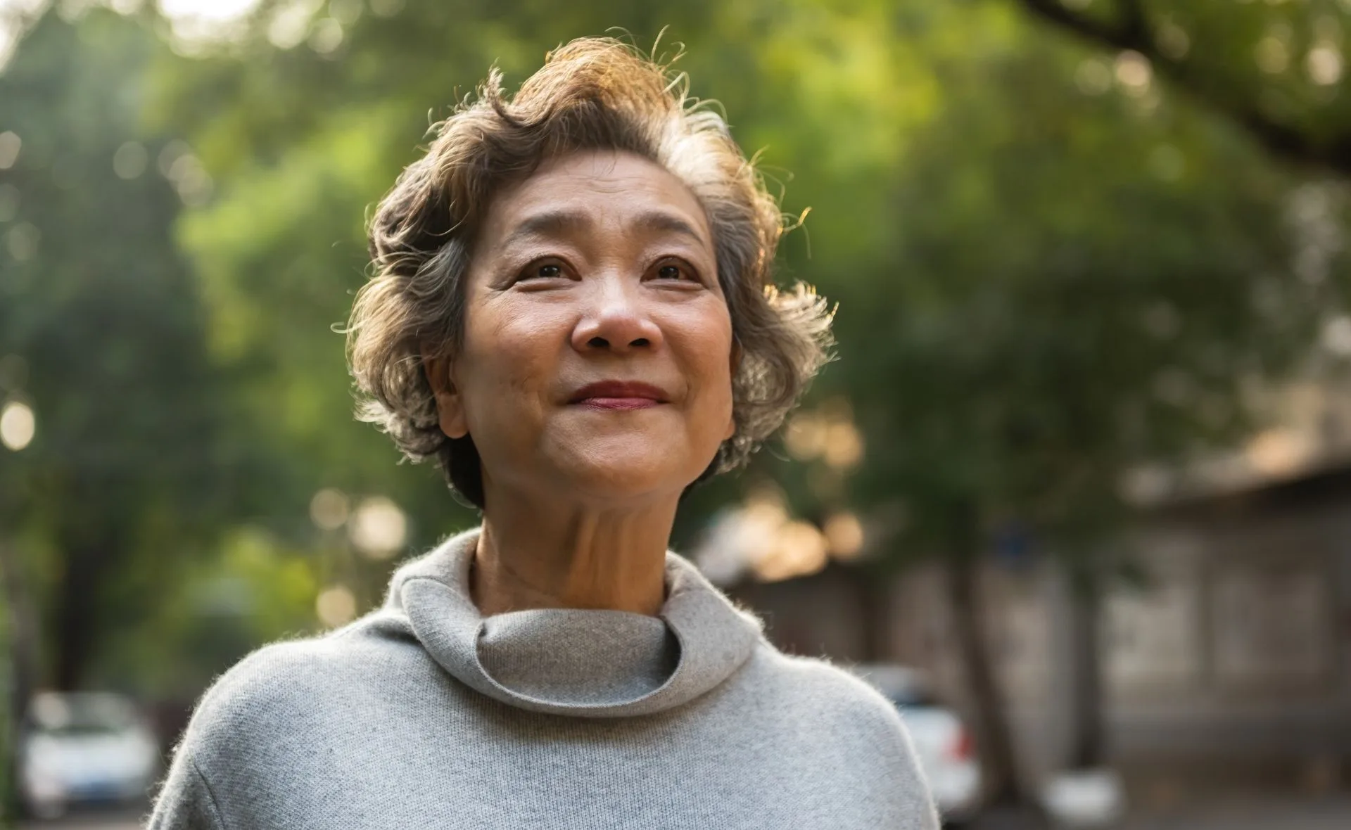 Senior woman with short hair smiling outdoors, wearing a gray sweater, with a soft green background.