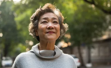 Senior woman with short hair smiling outdoors, wearing a gray sweater, with a soft green background.