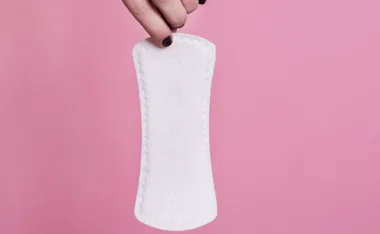 Hand holding a white biodegradable menstrual pad against a pink background.