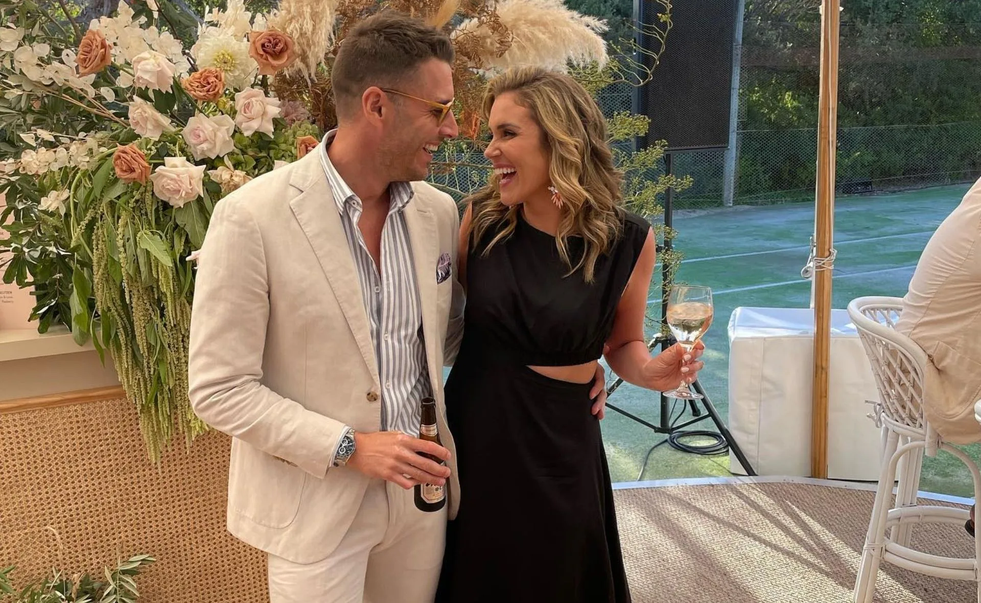 Couple smiling at each other, dressed elegantly, holding drinks, with floral decor and outdoor setting in the background.