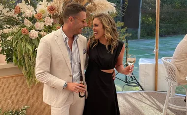 Couple smiling at each other, dressed elegantly, holding drinks, with floral decor and outdoor setting in the background.