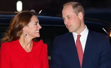 A smiling couple, the woman in a red dress and the man in a dark suit, stand together outside near a car.