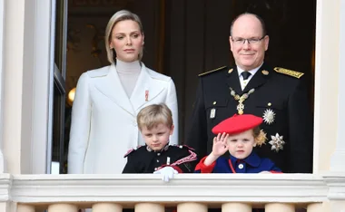 A family, including a woman in white and a man in black uniform with medals, stands on a balcony with two children.