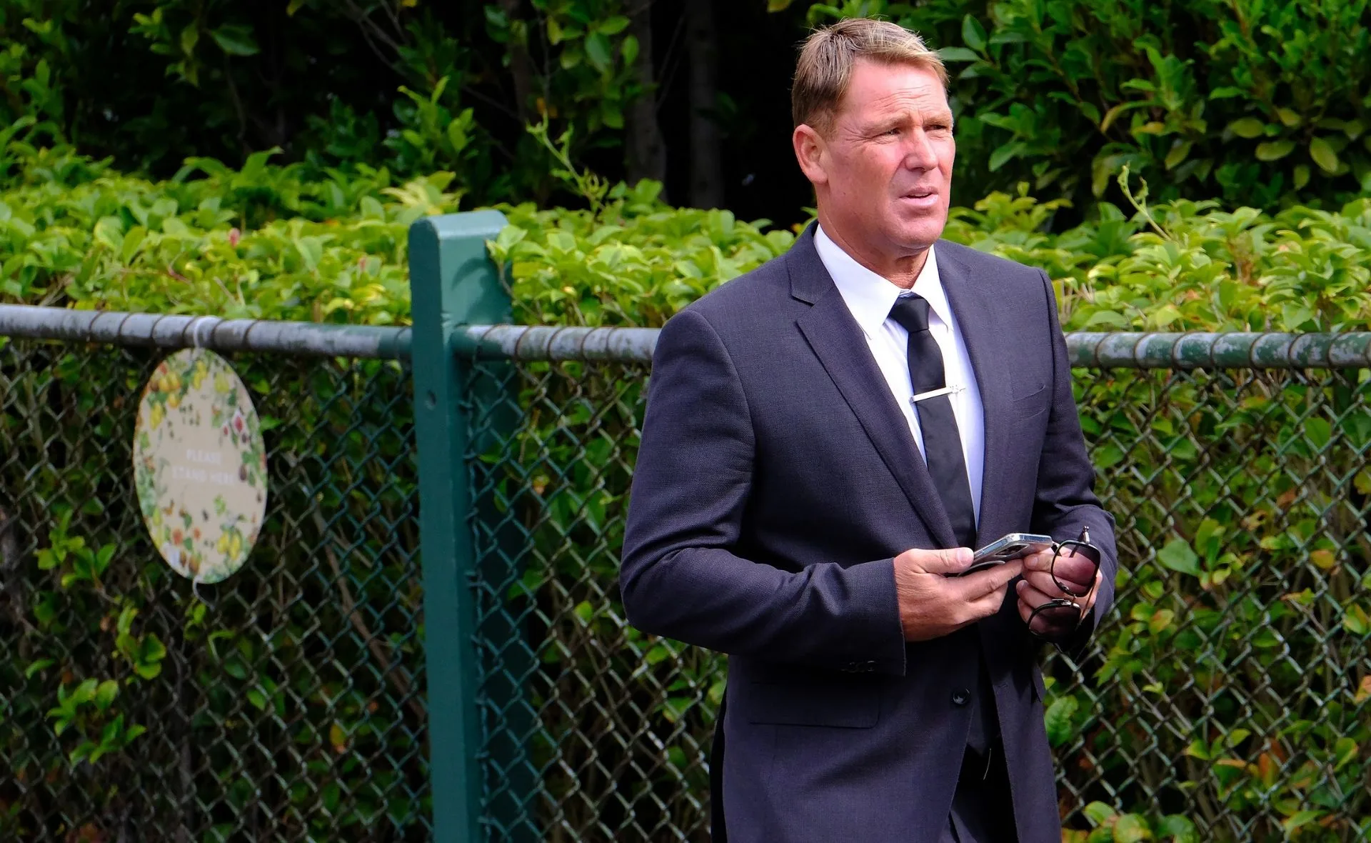 Man in a suit holding phone and glasses at a fence with greenery in the background.