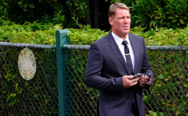 Man in a suit holding phone and glasses at a fence with greenery in the background.
