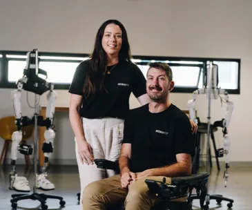 Young woman stands beside a man in a wheelchair; both wearing "Robotit" shirts with robotic exoskeletons in background.