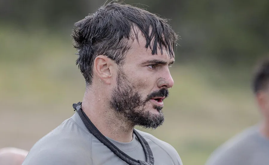 A man with short hair and a beard, looking focused, wearing a gray shirt and a rope around his neck outdoors.