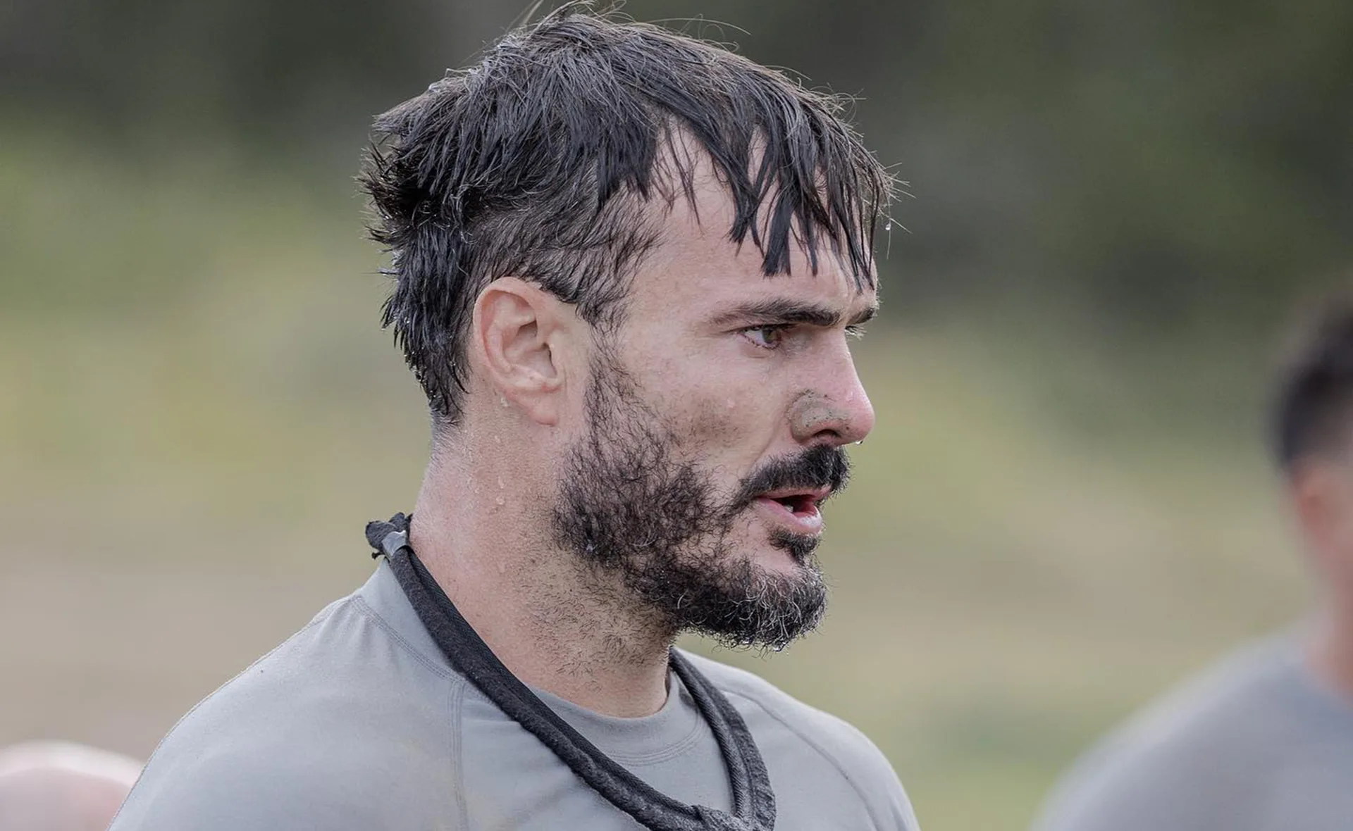 A man with short hair and a beard, looking focused, wearing a gray shirt and a rope around his neck outdoors.