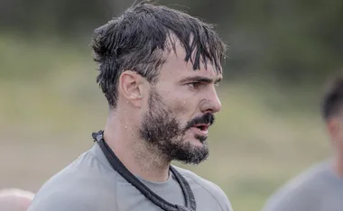 A man with short hair and a beard, looking focused, wearing a gray shirt and a rope around his neck outdoors.