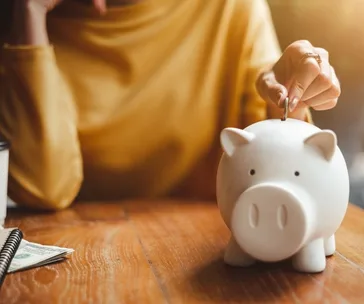 Person placing a coin into a white piggy bank on a wooden table, with a notebook and cash nearby.