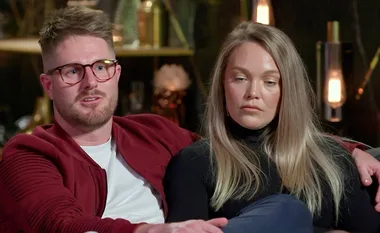 A man in glasses wearing a red jacket sits on a couch with a woman in a black top, both with a serious expression.