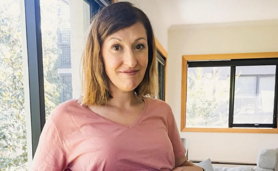 Woman in a pink shirt smiling indoors near a large window with greenery outside.