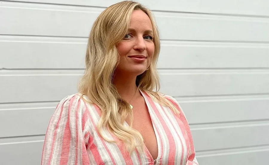 A woman with long blond hair in a pink and white striped dress smiles against a white paneled background.
