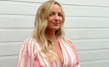 A woman with long blond hair in a pink and white striped dress smiles against a white paneled background.