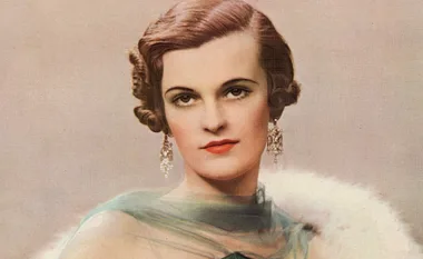 A woman with styled brown hair, wearing earrings and a fur stole over a sheer dress, posing in a vintage portrait.