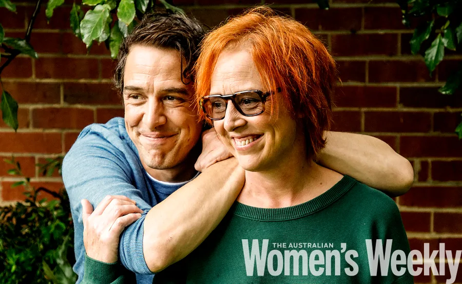 Man embraces woman outdoors, both smiling warmly. She wears glasses, both appear happy and content.