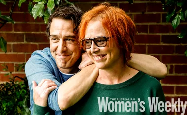 Man embraces woman outdoors, both smiling warmly. She wears glasses, both appear happy and content.
