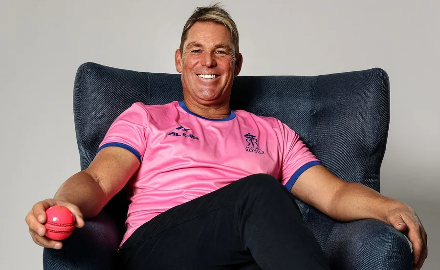 Man smiling, sitting on a chair, wearing a pink Rajasthan Royals shirt, holding a cricket ball.